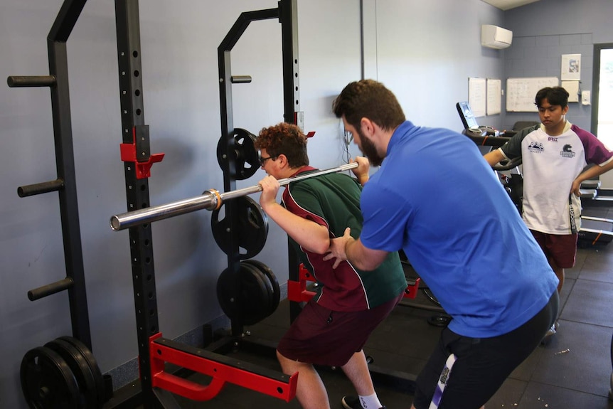 A teenager lifting weights in a gym with the help of an instructor
