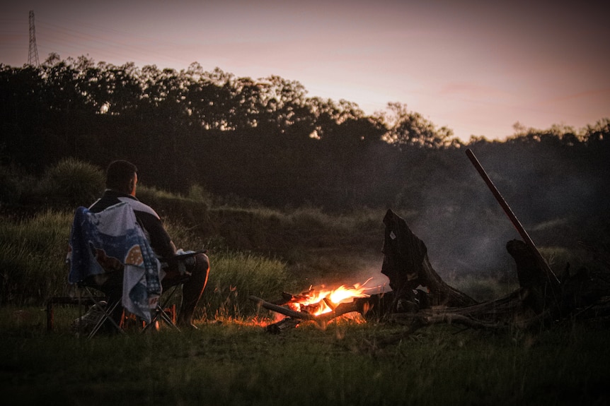 Man sitting in a camping chair by a small campfire as the sun sets.