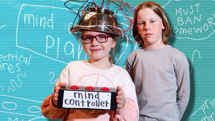A child holds a pretend mind control device and colander with wires attached on her head. Another child looks unimpressed.