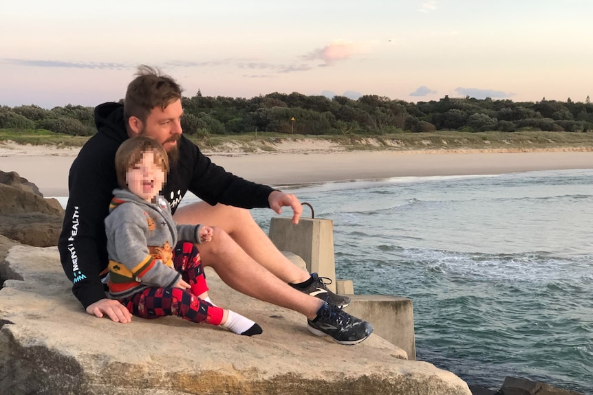 Dad in black jumper with his arm around son on a rock near a beach.