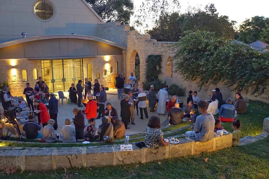 A group of people - some wearing Muslin head coverings - congregate in a church courtyard, eating and talking.