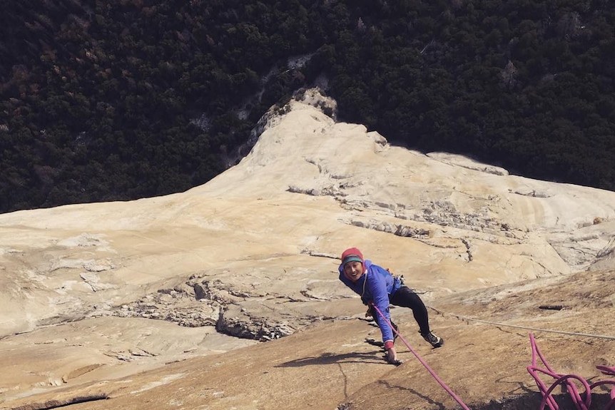 A rock climber in purple jacket using a rope to climb a steep rock face.