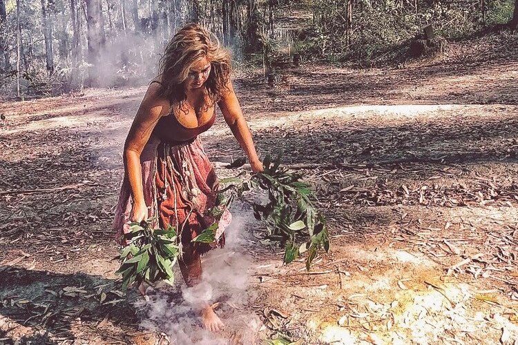 An indigenous woman dances next to a smoking fire holding gum leaves.