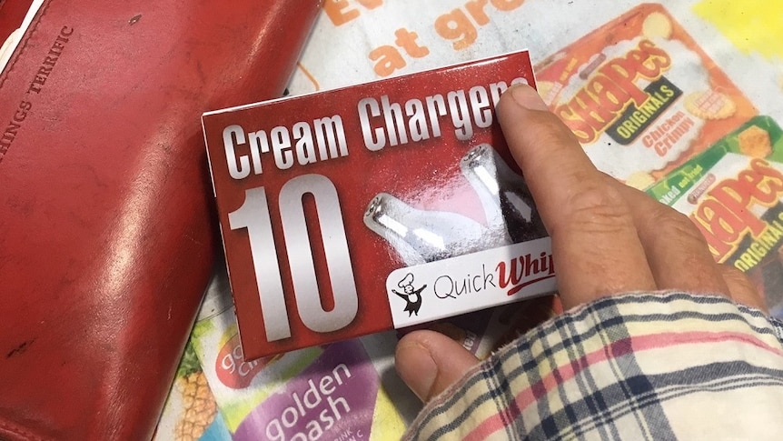 A hand holding a red packet of 'cream chargers' over a shop counter with a red wallet nearby