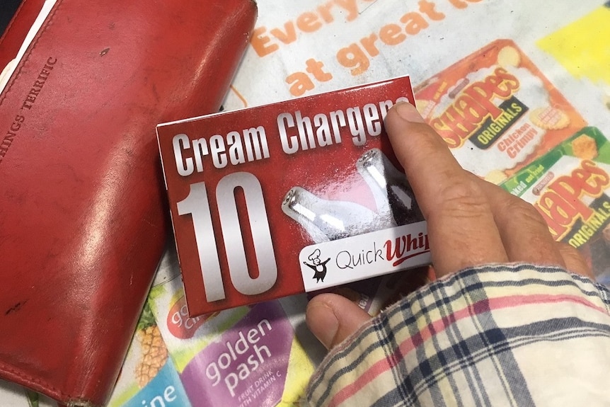 A hand holding a red packet of 'cream chargers' over a shop counter with a red wallet nearby