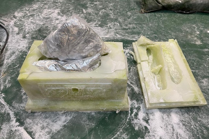 A foil packet protrudes from pale yellow-green stone-like box, with white powder surrounding it. The box is Oriental in design.