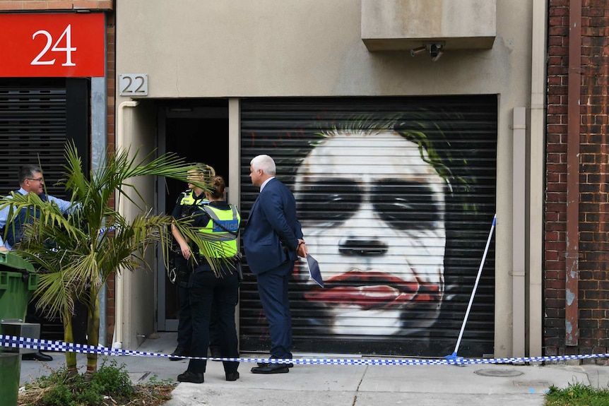 Police tape and four police officers can be seen outside a building with a painting of the Joker on a roller door.