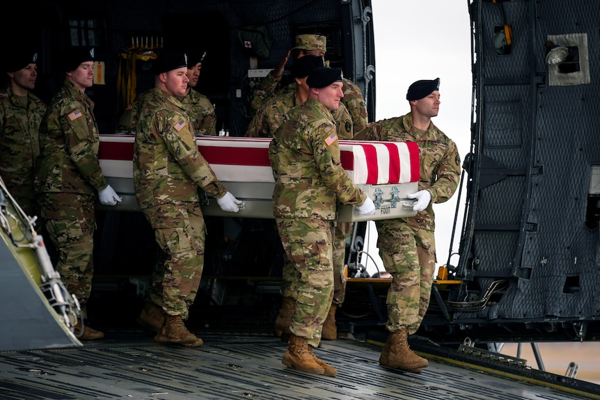 A group of US soldiers in camouflage uniform carry a flag-draped metal case out of an airplane.