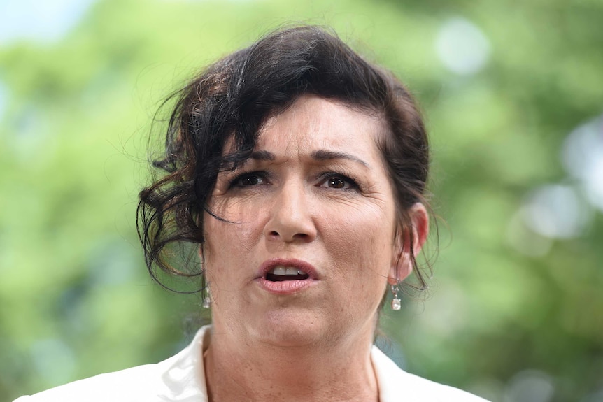 Head shot of a woman in a white jackets speaking at an outdoor press conference.