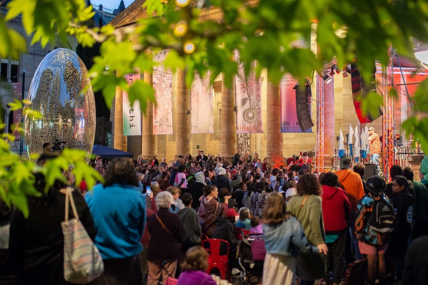 An evening shot of the packed entrance to the AGSA, lit up in colourful lights. People watch a performance.