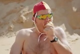 man in speedos eating an onion on the beach