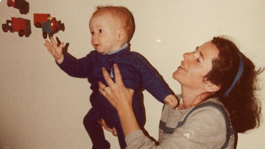 Young woman holding a baby son up to a chidren's truck mobile. Mother is smiling. Baby's arm is reaching up to touch the mobil