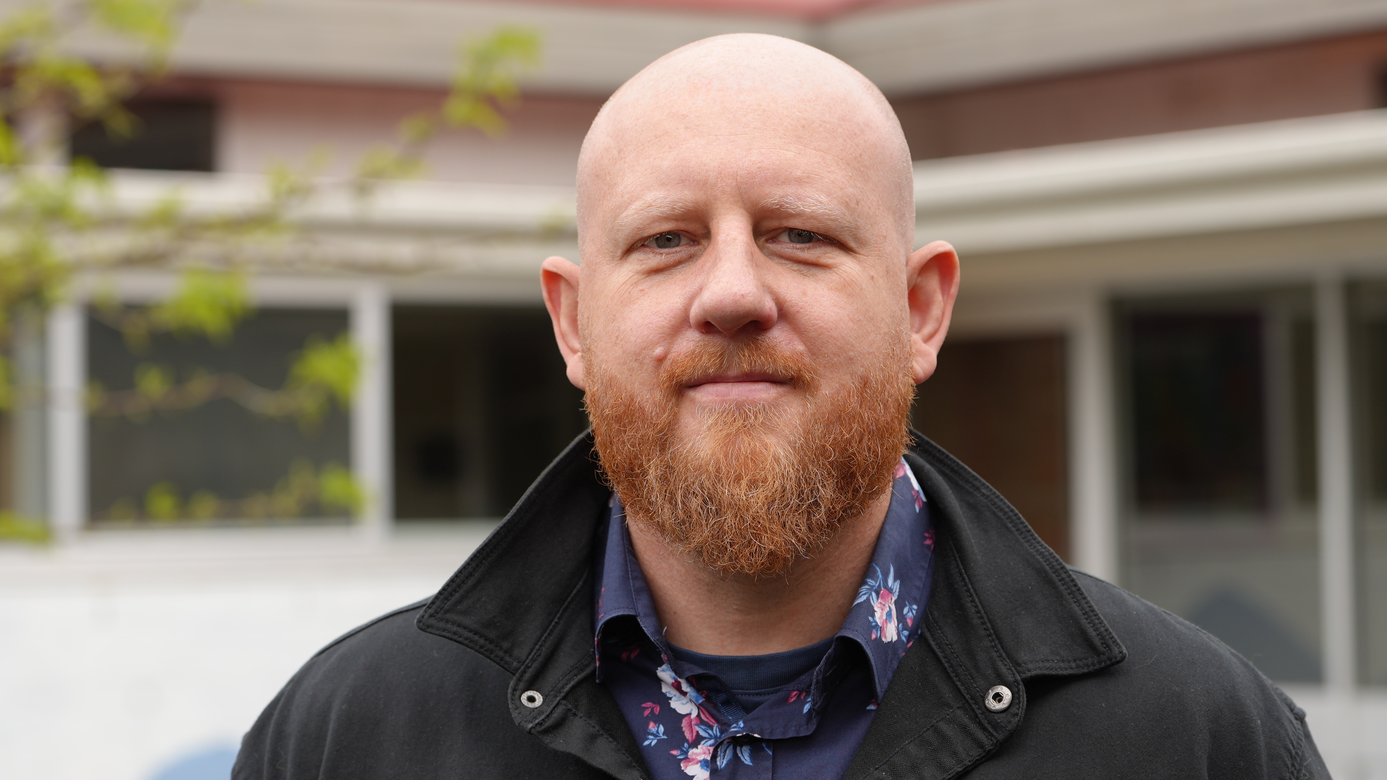 A bald man with a red beard stands outside a building looking serious.