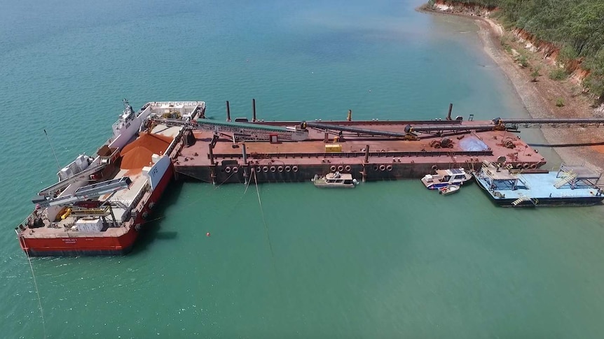 Large red piles of bauxite are visible on the open deck of a barge being loaded at Hey Point, near Weipa