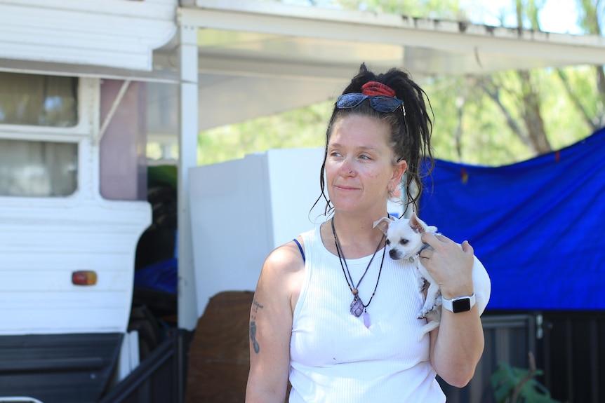 A woman with long, dark hair stands in front of a caravan, cradling a small dog.
