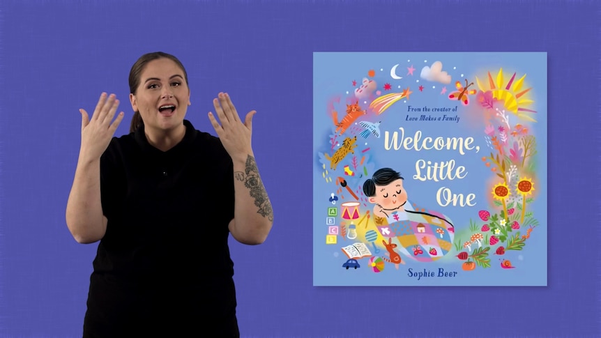 Auslan presenter Michelle Rowlands stands beside image of book, Welcome, Little One