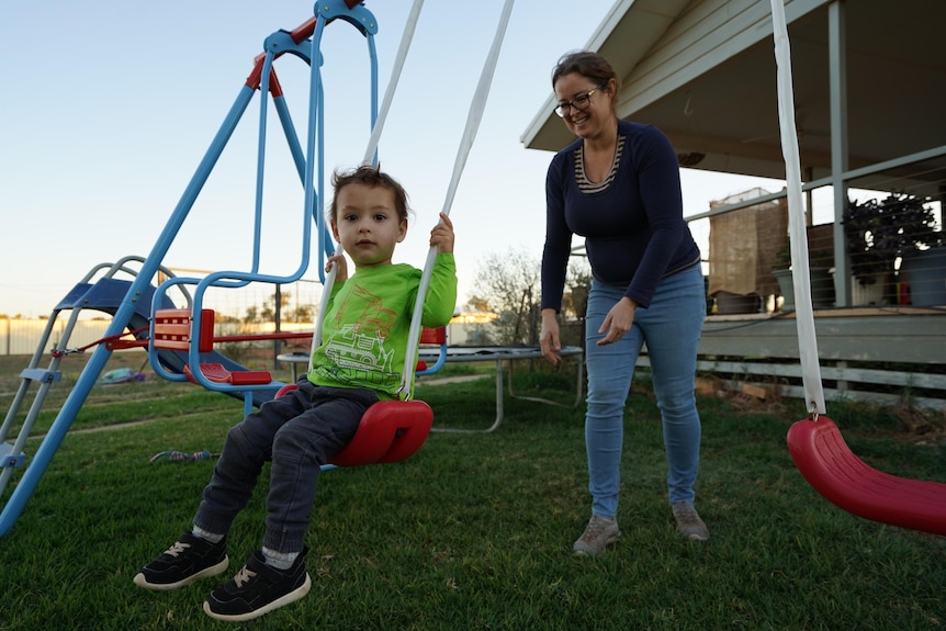 Woman pushes young boy on a swing set on lawn outside house.