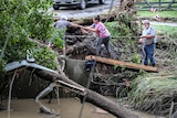 Three people holding a thin rope carefully cross ground next to floodwaters.