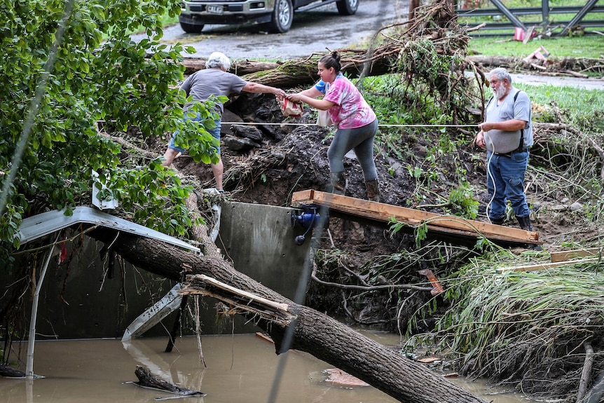 Three people holding a thin rope carefully cross ground next to floodwaters.