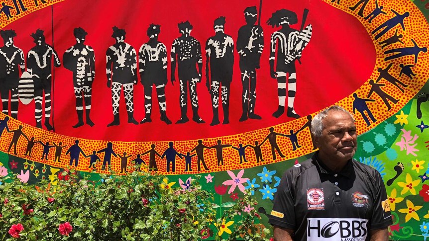 Aboriginal man stands respectfully, with hands clasped in front of him, as he stands in front of a colourful Indigenous mural