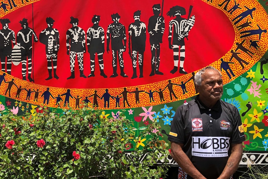 An Aboriginal man stands respectfully, with hands clasped in front of him, as he stands in front of a colourful Indigenous mural