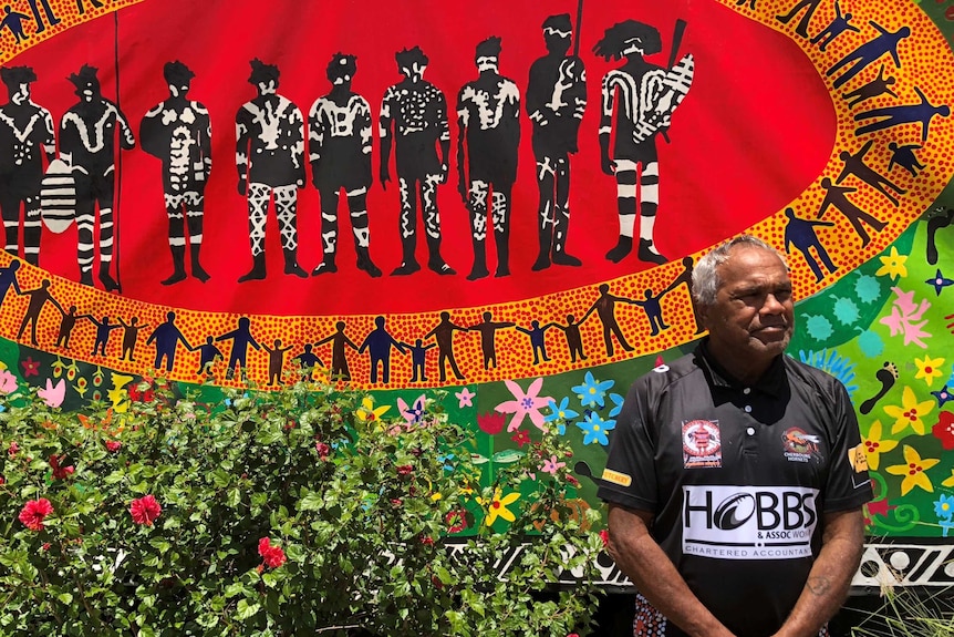 Aboriginal man stands respectfully, with hands clasped in front of him, as he stands in front of a colourful Indigenous mural