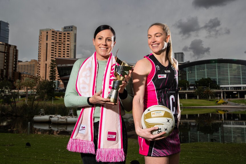 A former player holds a trophy and wears a pink scarf while a current player holds a netball, stares at trophy