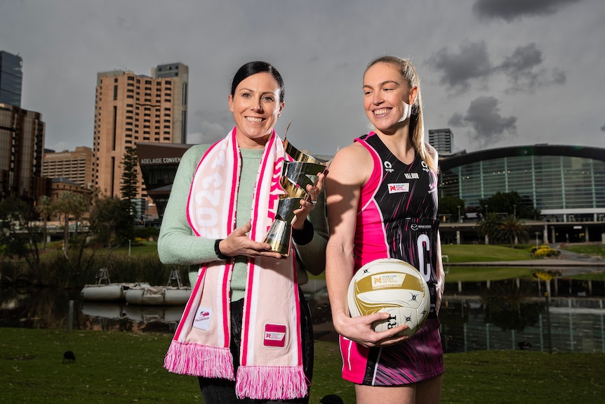 A former player holds a trophy and wears a pink scarf while a current player holds a netball, stares at trophy