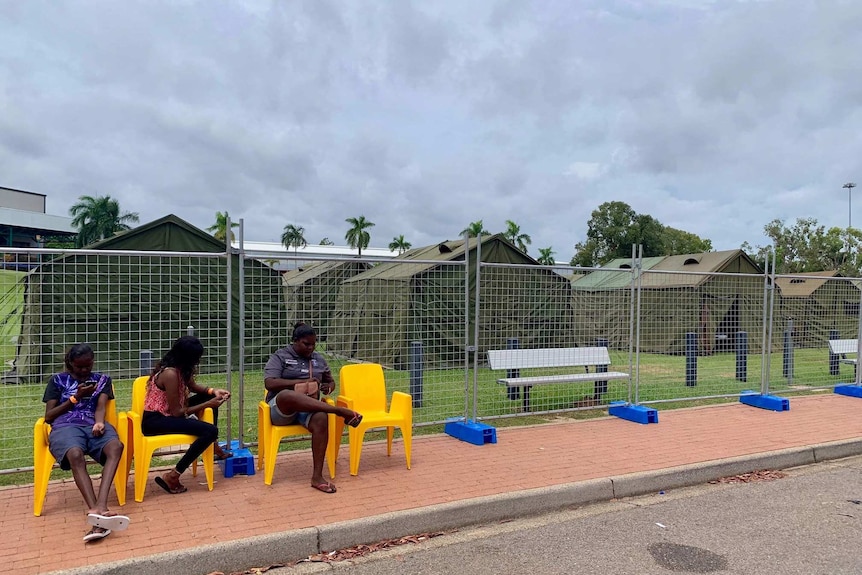 People sit on chairs in front of a fenced tent community.