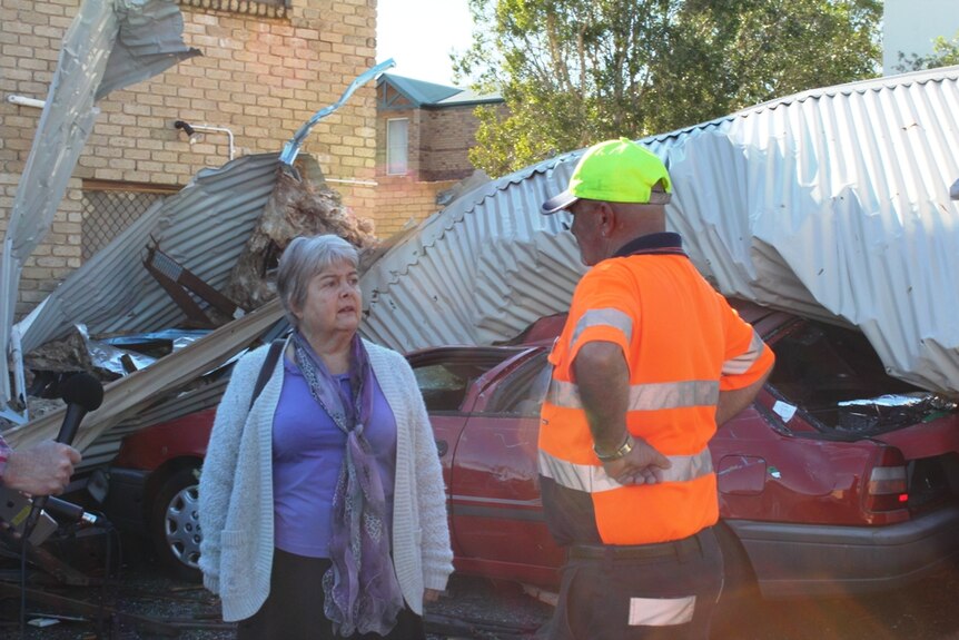 Elderly woman talking to a man wearing hi-vis and in front of car with tin roof smashed into it.