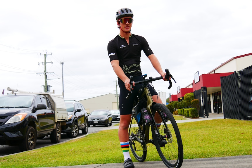 looking up at a young man on his bike in a lycra shirt and shorts with a helmet on, green grass, cars and buildings behind 