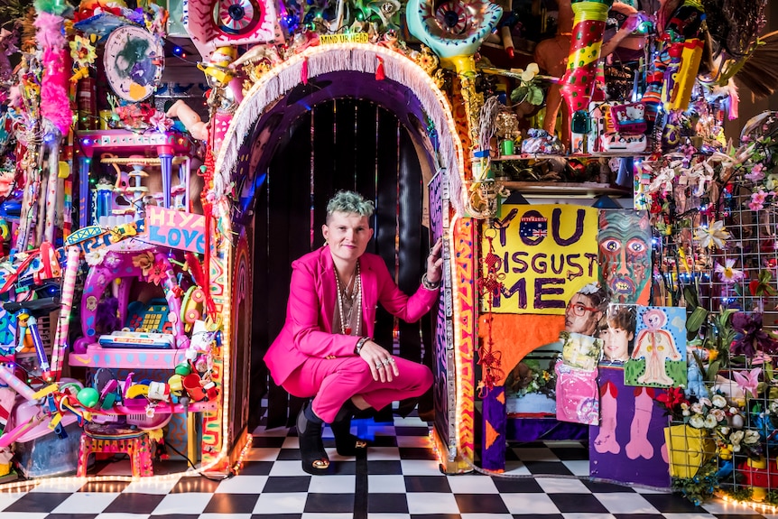 A young man with bleach blond hair wearing a hot pink suit crouches in front of his colourful artwork