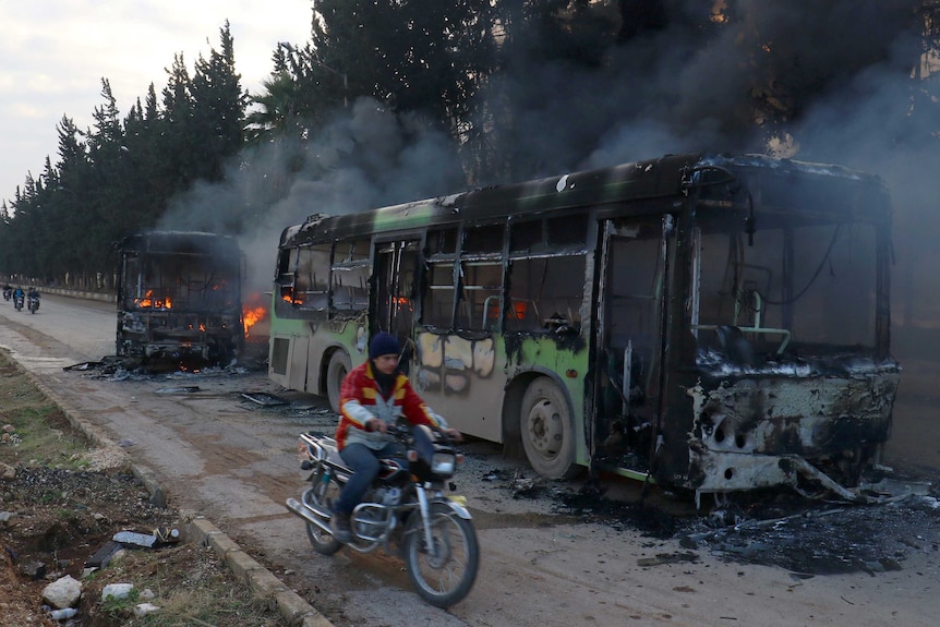 A man on a motorbike bike rides past burning buses.