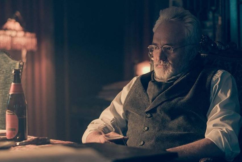 Bradley Whitford sitting in a dark room with a bottle of bear on the table in front of him.