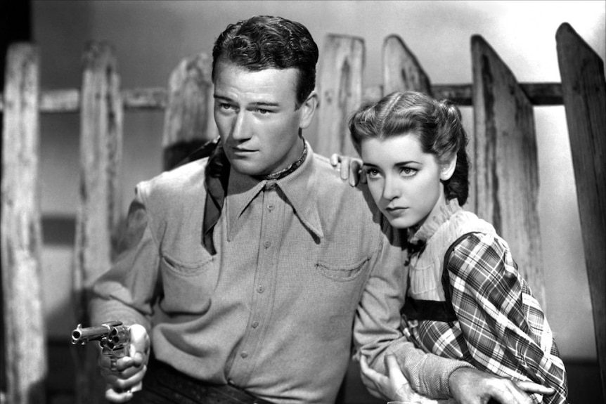 A screen grab from a black and white western movie, where a young Marsha hunt is hiding slightly behind a man with a gun.