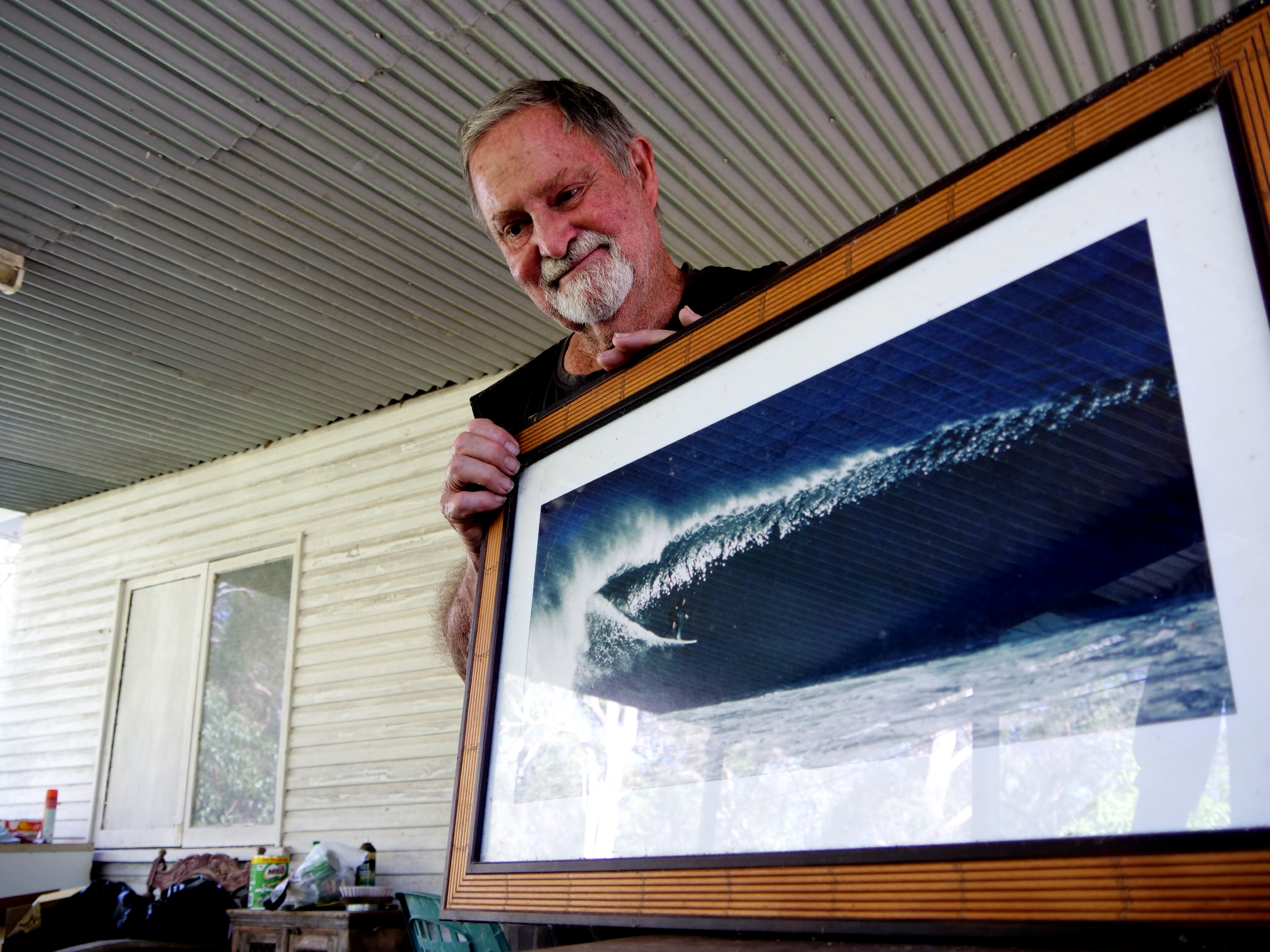 Un hombre mayor sostiene un marco de fotos con una gran ola que lo muestra surfeando cuando era joven