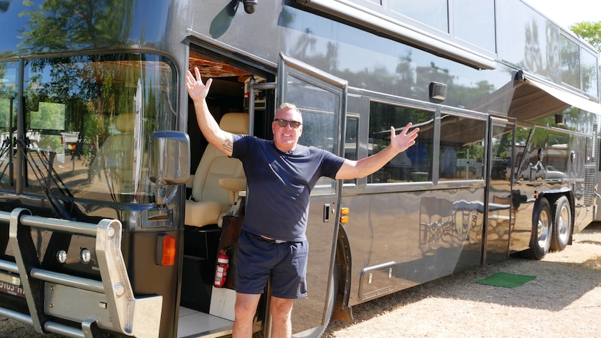 A man in dark shirt and shorts raises arms, smiling in front of a large black double-decker bus