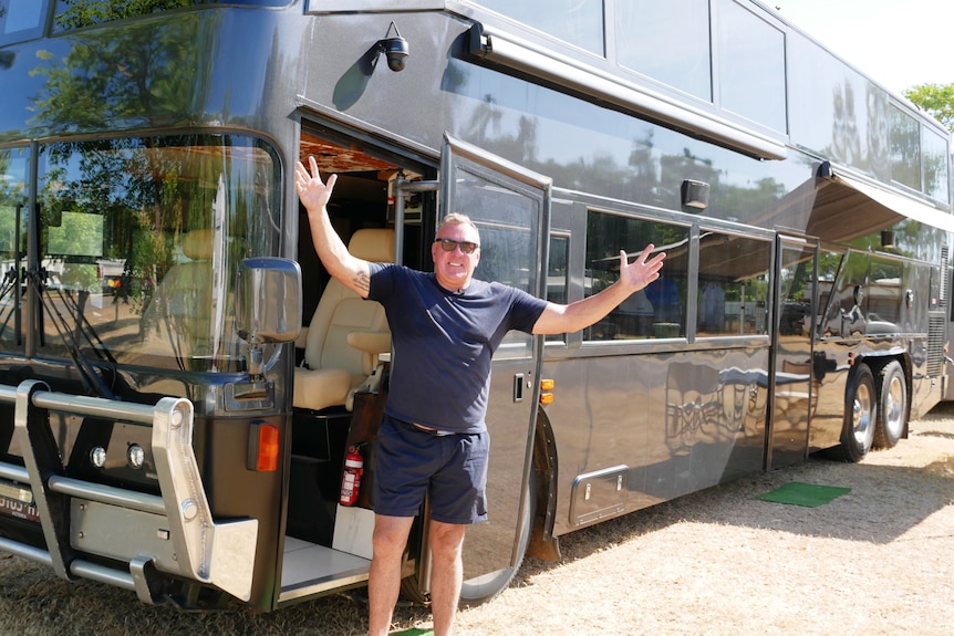 A man in dark shirt and shorts raises arms, smiling in front of a large black double-decker bus