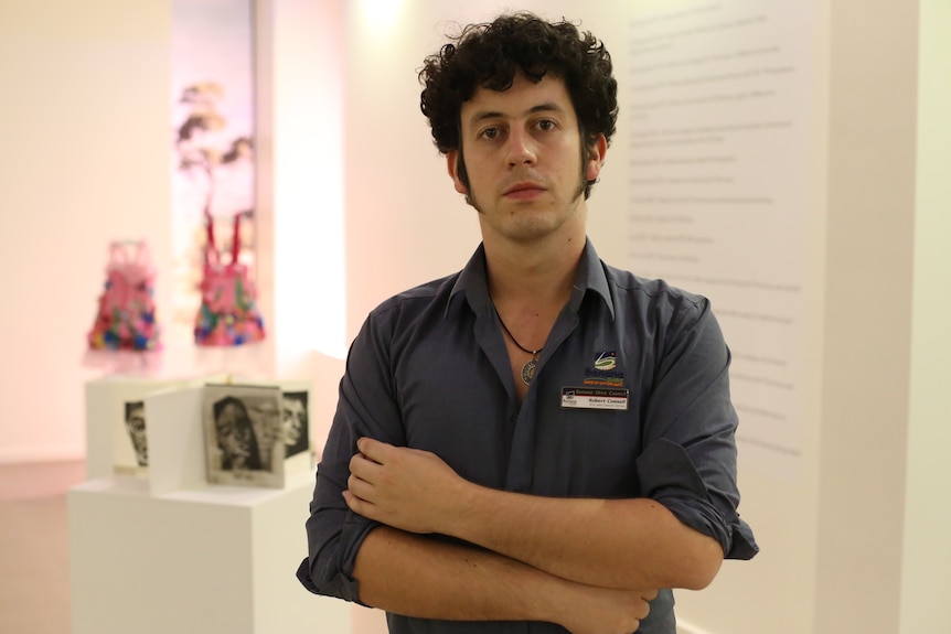 A serious man with curly dark hair, long sideburns, grey shirt, arms crossed, stands in front of displays in an art gallery.