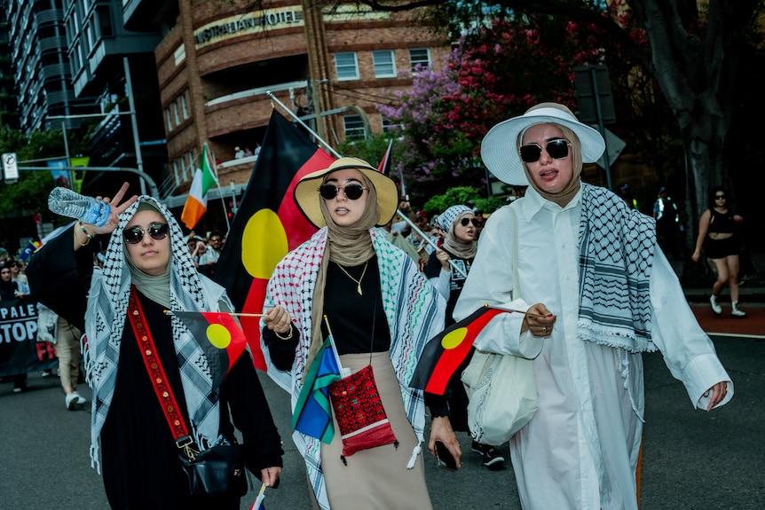 Three women wearing Palestinian scarfs march in an Invasion Day protest, holding Aboriginal flags