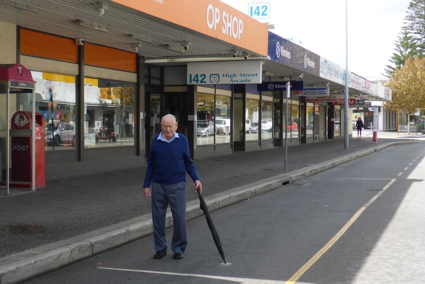An elderly man in a blue jumper points an umbrella at a spot on a road outside a shop front.