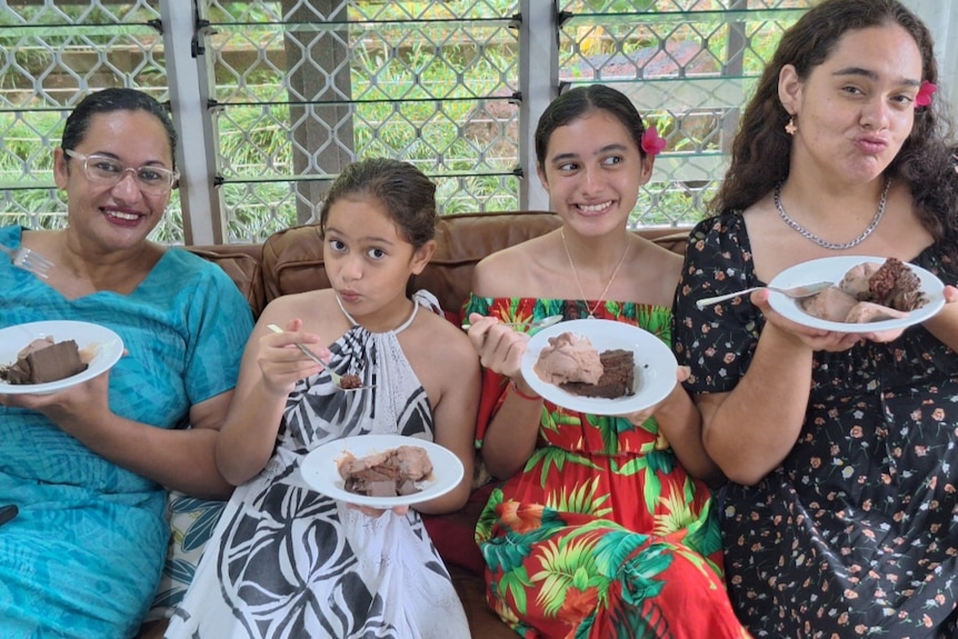 A mother and three daughters with colourful floral dresses, holding plates of food and smiling. 