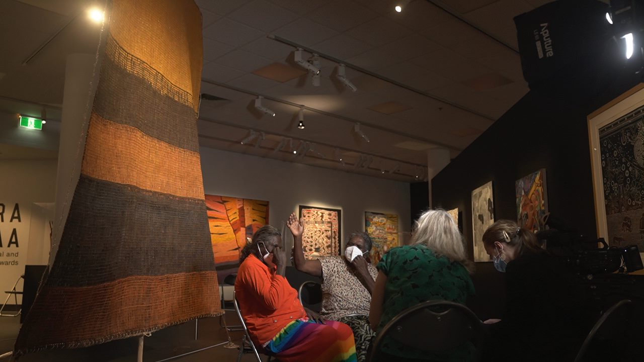 Women sitting beside a large woven artwork.