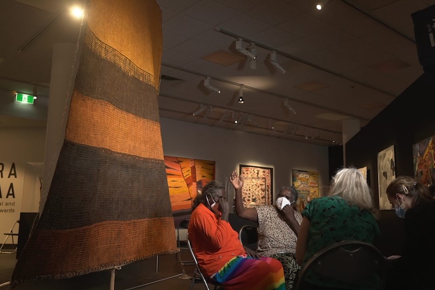 Women sit next to a large woven artwork.