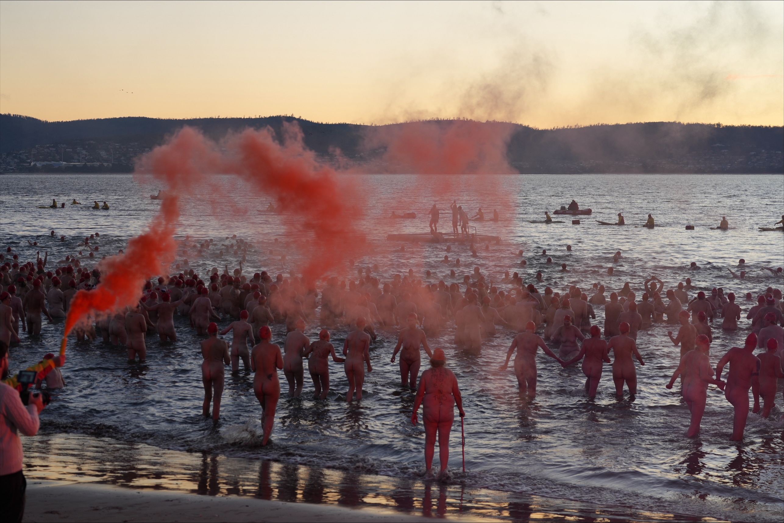 a crowd of naked people run into the river as red flares are shot into the air.
