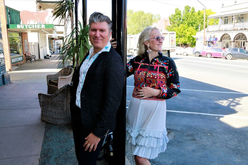 Couple from town of Braidwood stand at pole on town street, with man looking at camera while woman smiling towards street.