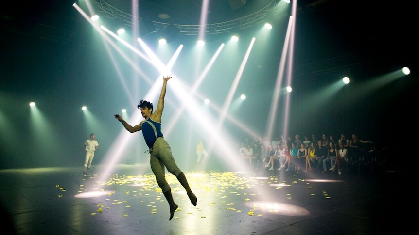 A darkened elliptical stage and a spaceship-like lighting rig suspended from  ceiling and a dancer in athletic gear mid-starjump