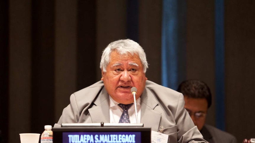 Prime Minister Tuilaepa Lupesoliai Sailele Malielegaoi sits behind a birch desk and his name card as he speaks into a microphone