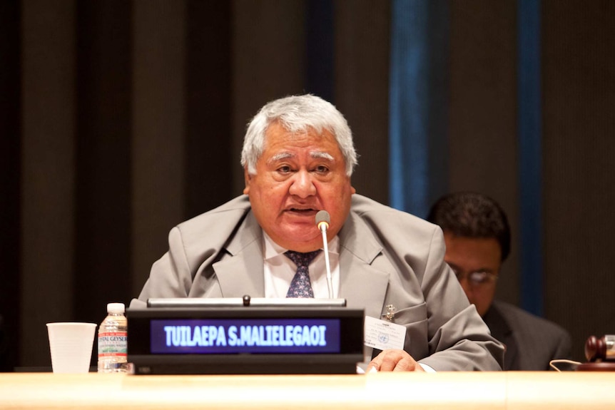 Prime Minister Tuilaepa Lupesoliai Sailele Malielegaoi sits behind a birch desk and his name card as he speaks into a microphone