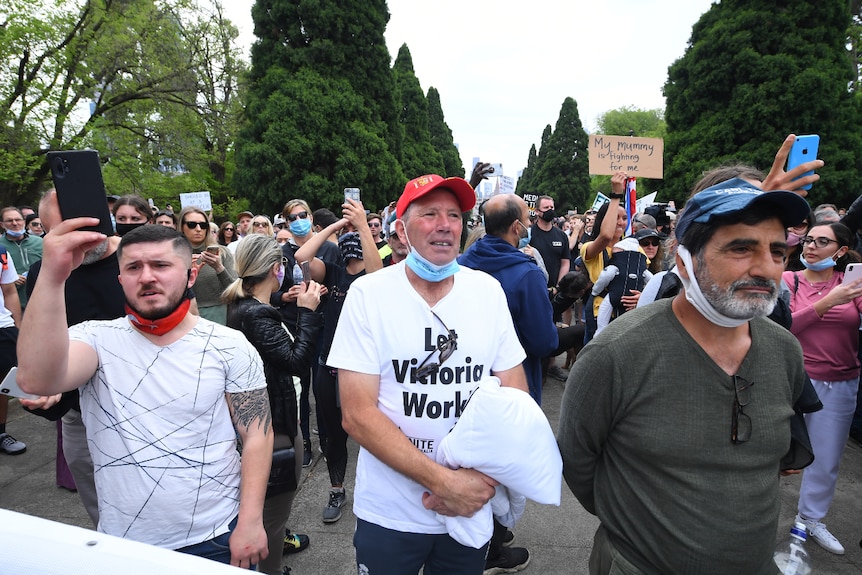 Three men with face masks below their chins stand in a crowd.